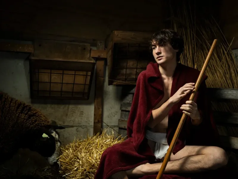 Artwork JON THE BAPTIST by Arjan Spannenburg showing a young man draped in a red cloak holding a wooden staff in a dark stable with a sheep.