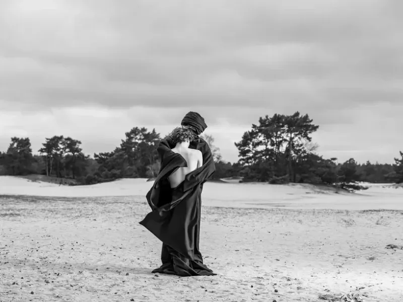 A high-contrast monochrome photograph by Arjan Spannenburg titled "EMERGE." The composition focuses on two figures in a desolate sandy landscape. A man with his bare back to the camera finds sanctuary in the dark, layered robes of a second figure, whose head is wrapped in a traditional covering. The wind catches the fabric against a minimalist horizon.