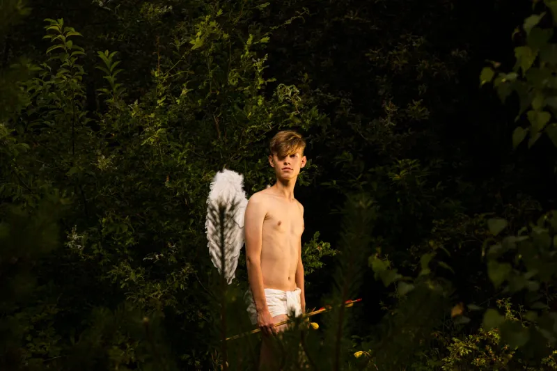 A young man with wings and a bow stands among dark leaves, embodying a vulnerable contemporary Cupid.