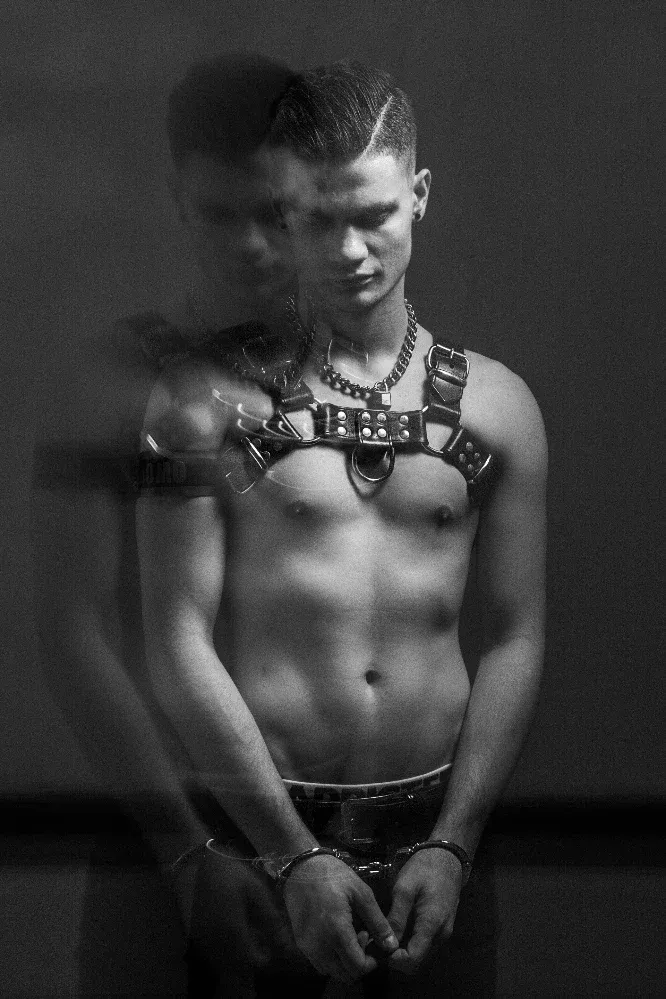 A high-contrast black and white photograph by Arjan Spannenburg titled WORRIED, featuring a man in a leather harness and handcuffs with a downward gaze and a blurred secondary exposure.