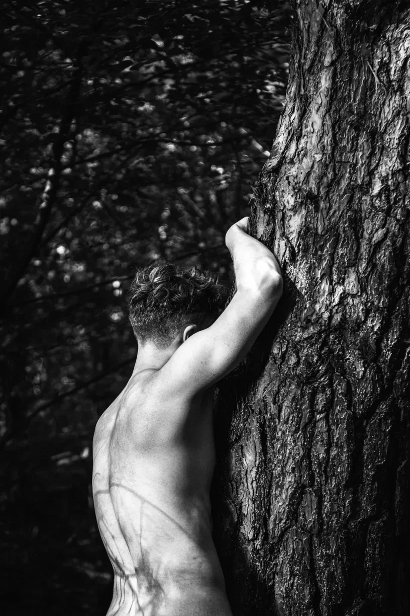 A black and white fine art photograph capturing a masculine back leaning into a textured tree trunk, emphasizing skin texture and shadows of foliage to represent masculine vulnerability and a connection to nature.