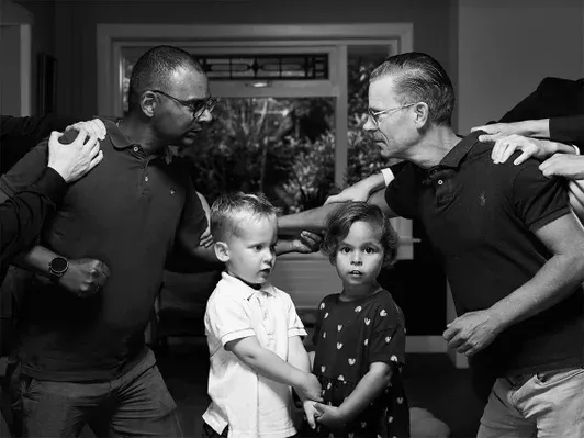 Black and white conceptual fine art photograph 'SANCTIONED' by Arjan Spannenburg, featuring a family in a protective stance while several hands reach in from the edges to pull them apart, highlighting themes of resilience and social pressure.