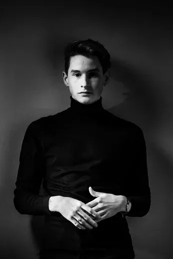 Fine art black and white portrait titled 'The Observer' by Arjan Spannenburg. A young man in a black turtleneck stares intently at the viewer with clasped hands. Winner of the FEP Awards 2019 Merit in Portraiture.