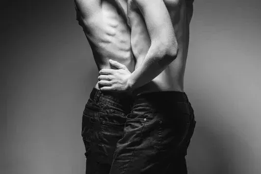 A high contrast black and white photograph of two men embracing, focusing on the waist and hands, exploring themes of intimacy and velleity.